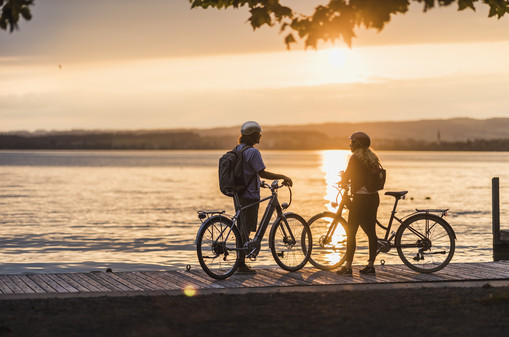Velofahrer am Seeufer des Zugersees bei Sonnenuntergang Zwei Velofahrer verweilen am Seeufer bei Sonnenuntergangsstimmung
