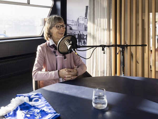 Eliane Müller sitzend am Tisch mit blauem Schal im Vordergrund in Podcast Mikrophon sprechend