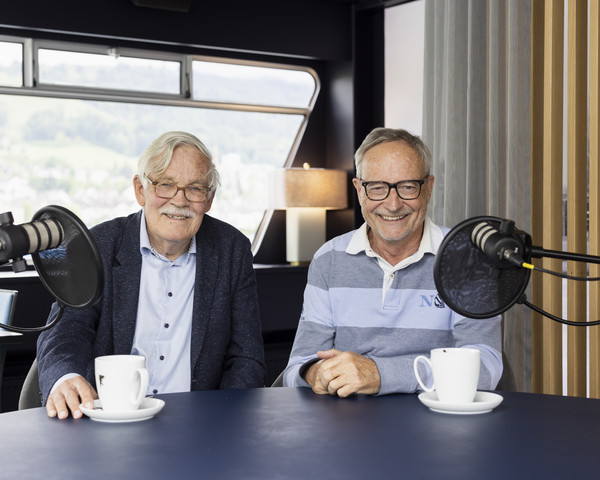 Jürg Meister und Heinz Amstad im Porträt am Podcast-Tisch sitzend vor den Mikrophonen