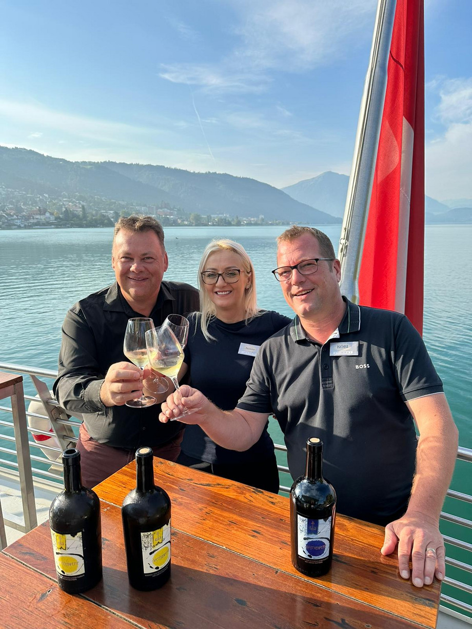 Anstossen auf den eigenen Wein Drei Personen stossen mit Gastronautic-Wein an, im Hintergrund ist der See